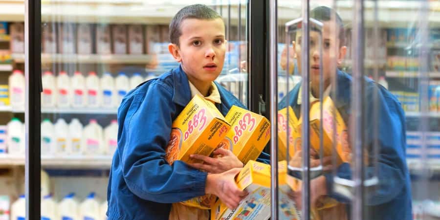 The image shows a young person, possibly a child, wearing a blue jacket and holding several boxes of what appears to be a food product in a grocery store aisle.